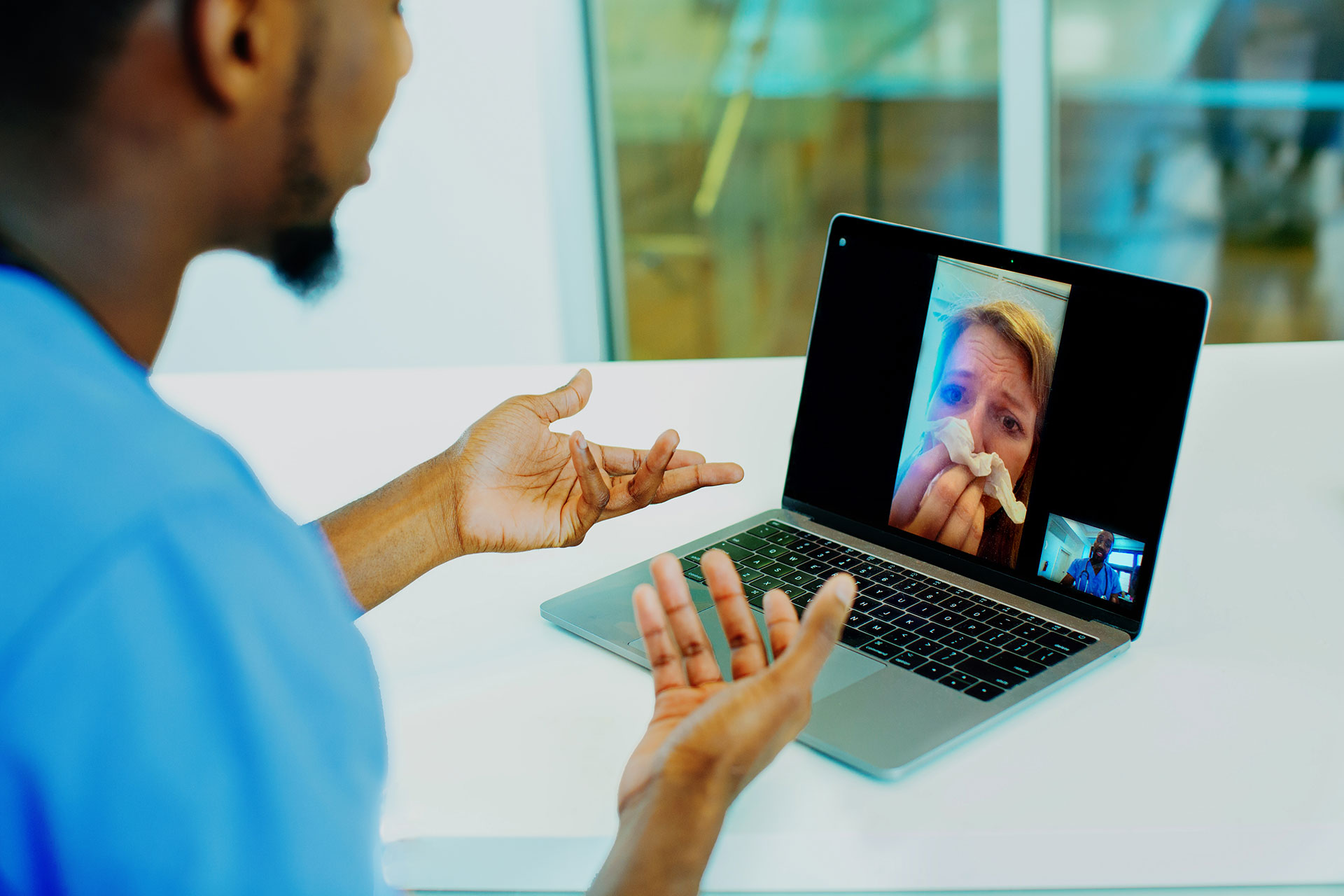 Portrait of a sick patient coughing into tissue being helped via tele medicine by a male doctor wearing blue scrubs uniform using laptop